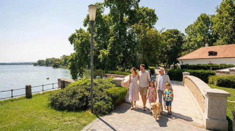 A képen egy család sétál a tatai Malomkert környékén, rendezett vízparti sétányon, zöld környezetben, háttérben az Öreg-tó hangulatával. Ez a tatai Malomkert séta helyszín kellemes választás azoknak, akik nyugodt, könnyen bejárható tatai programot keresnek családdal vagy akár kutyával. A vízparti séta és a közeli tatai látnivalók együtt igazán hangulatossá teszik a pihenést Tatán.